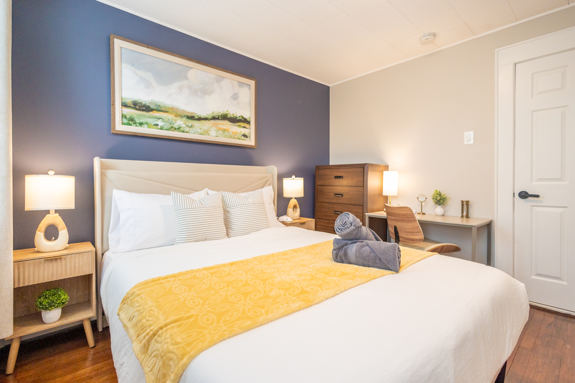 A bedroom with a queen bed, yellow blanket, blue accent wall, bedside tables with lamps, and a dresser with a desk.