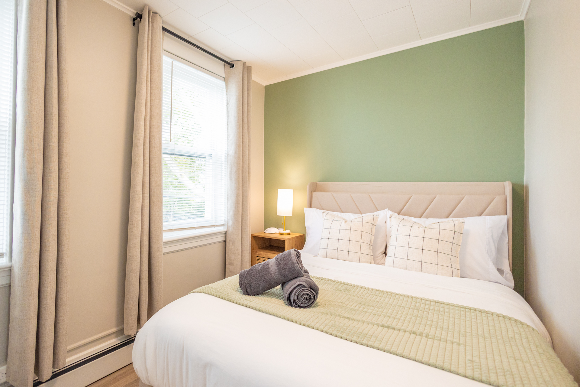 A bedroom with a double bed dressed in white linens and green accents, next to a nightstand with a lamp.
