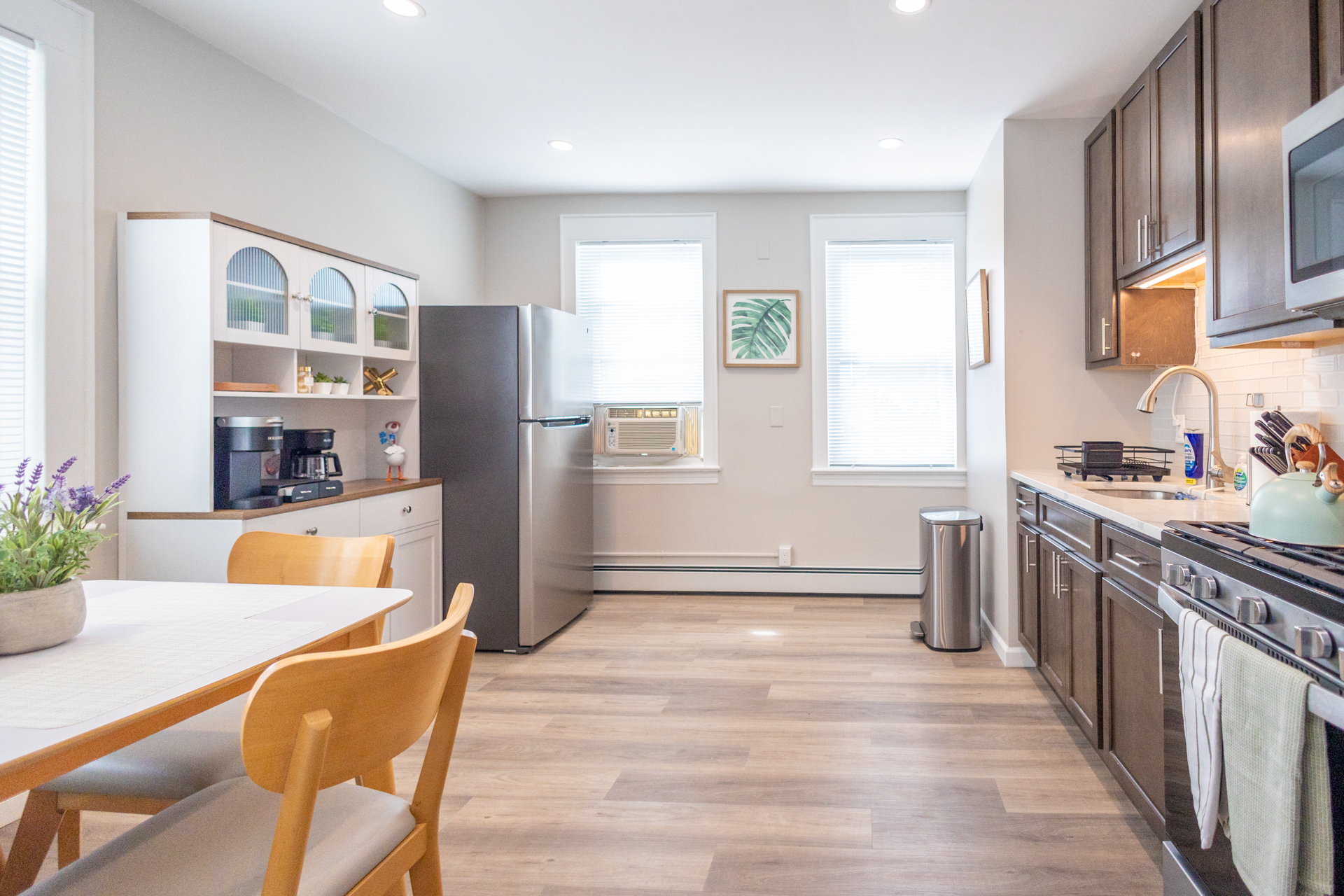 A modern kitchen with dark wood cabinets, stainless steel stove and microwave, and a dining table set for four. A white hutch with coffee station and fridge stand against the opposite wall.