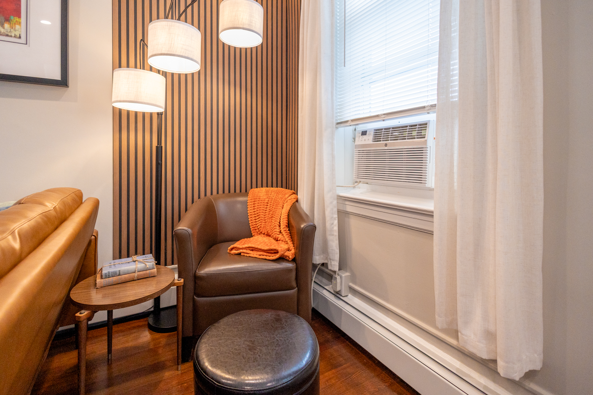 A cozy corner with a brown armchair, orange throw blanket, ottoman, side table with books, and a tall floor lamp.