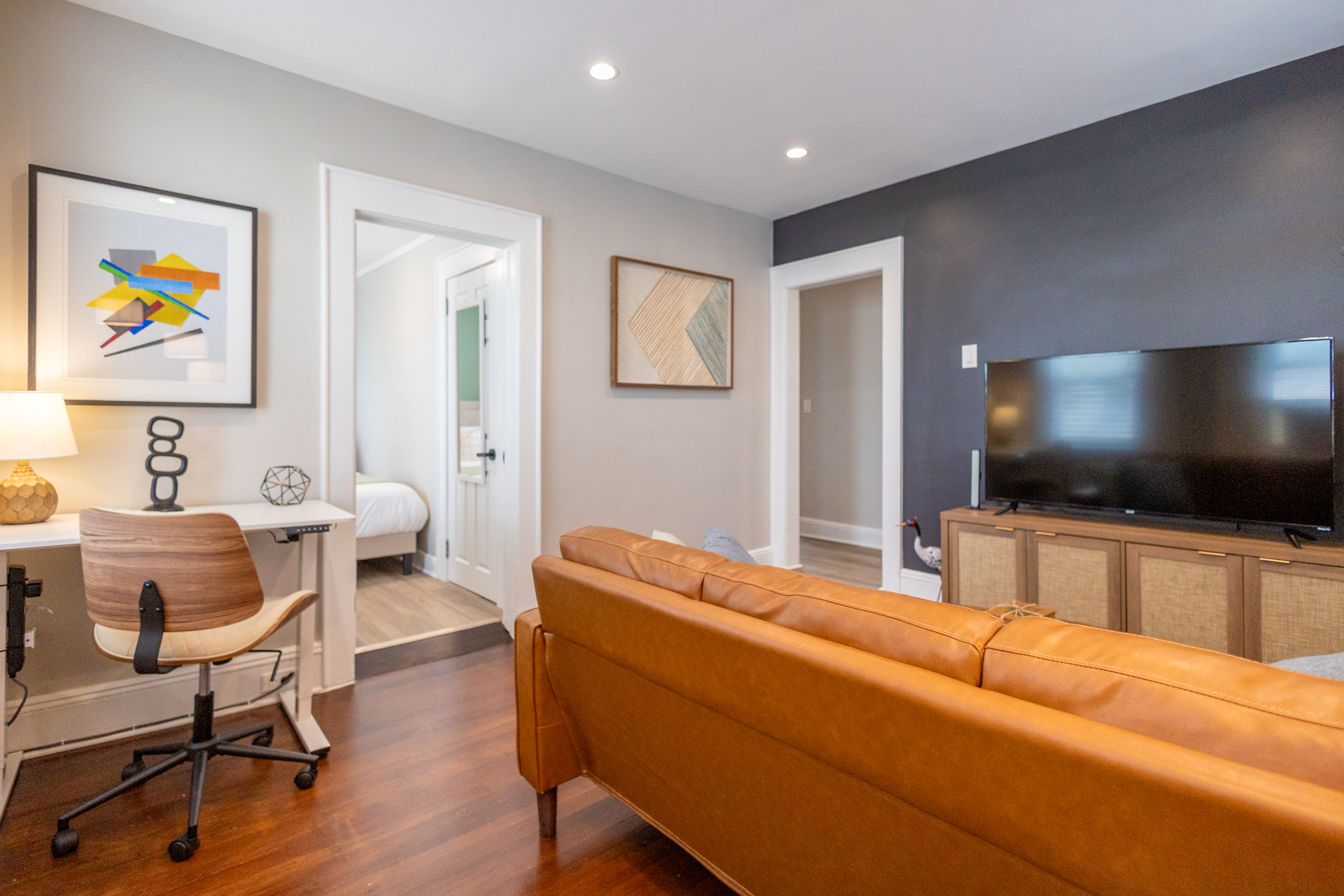 A living room with a leather sofa facing a TV on a wooden console. To the side, a small desk with a chair and lamp is tucked under framed art. The bedroom with green accent wall is visible through the doorway.