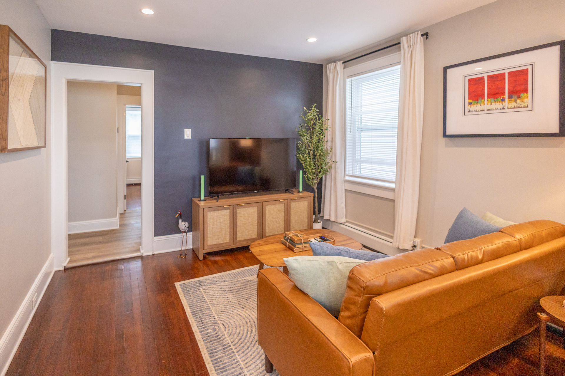 A TV on a wooden stand against a dark accent wall, with a leather sofa and patterned rug in the foreground. A window with white curtains sits beside the TV.