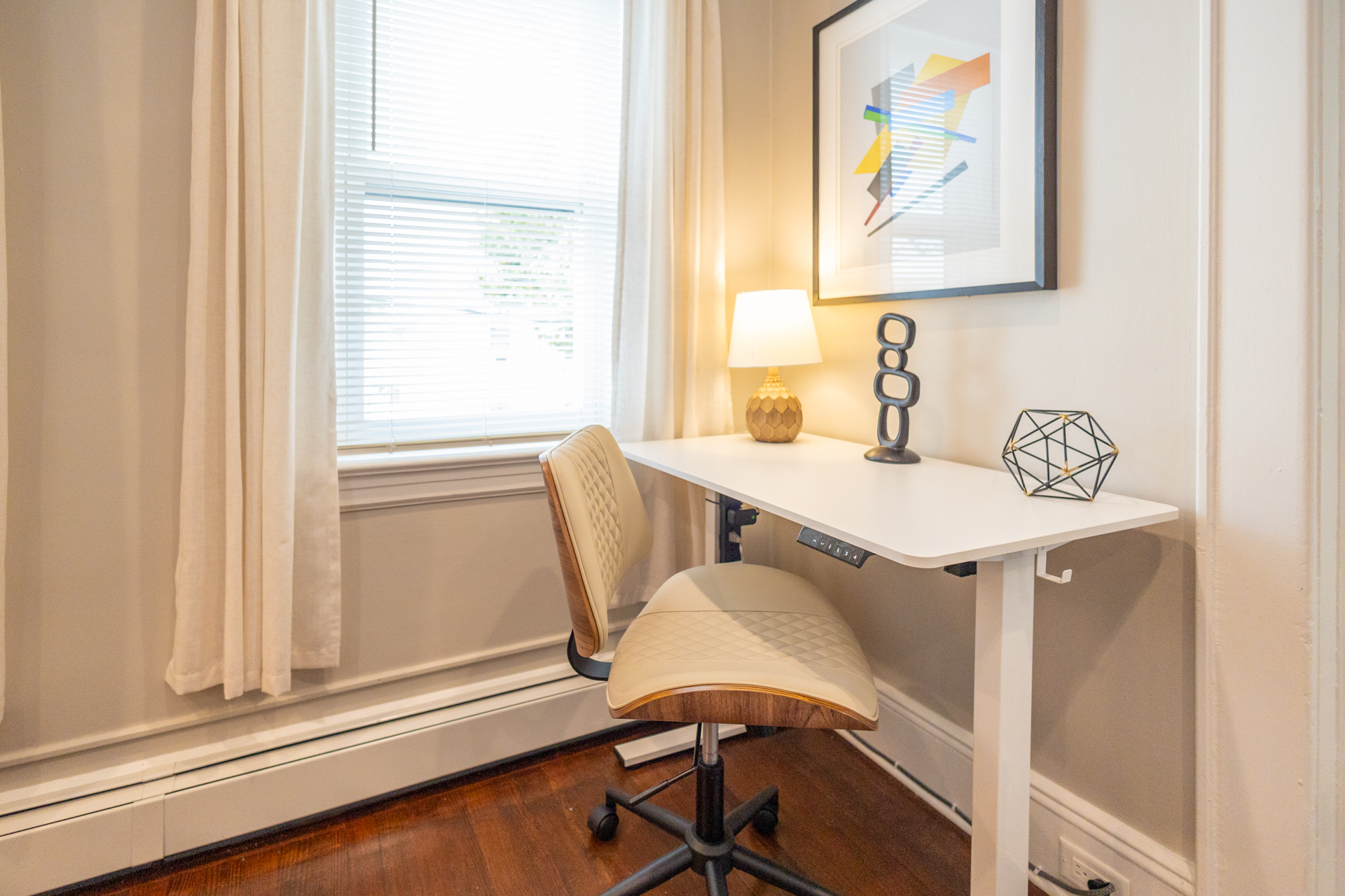 A small home office corner with a white desk, modern chair, lamp, and wall art near a window with curtains.