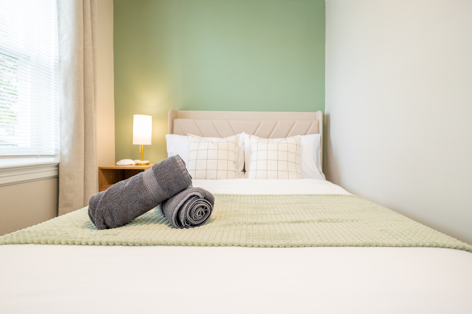 Straight-on view of a double bed with green accent blanket and pillows, with a wooden nightstand and lamp at the side.