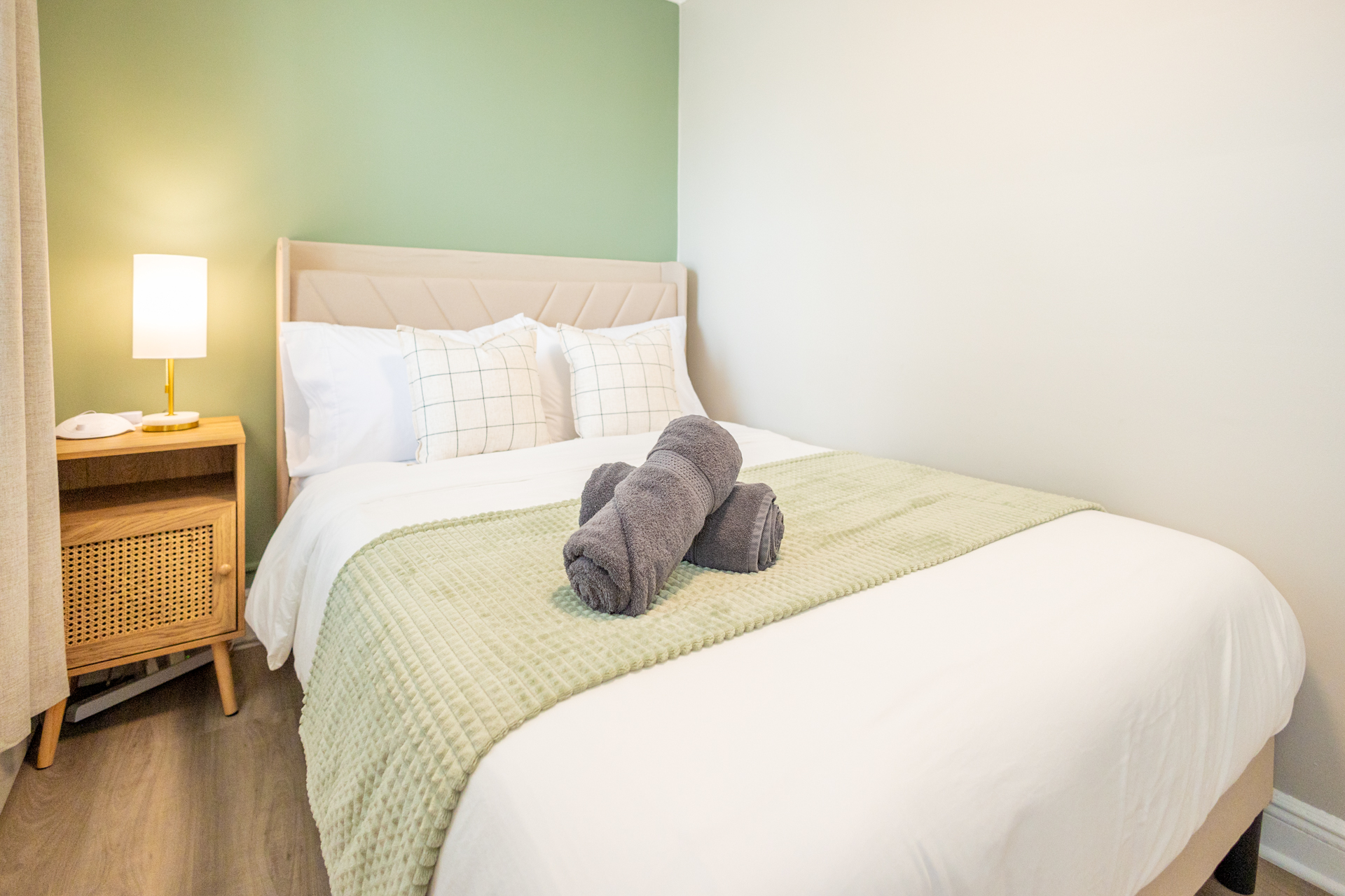 A double bed with white linens, green accent blanket, and plaid pillows against a soft green feature wall. Rolled gray towels rest on the bed.