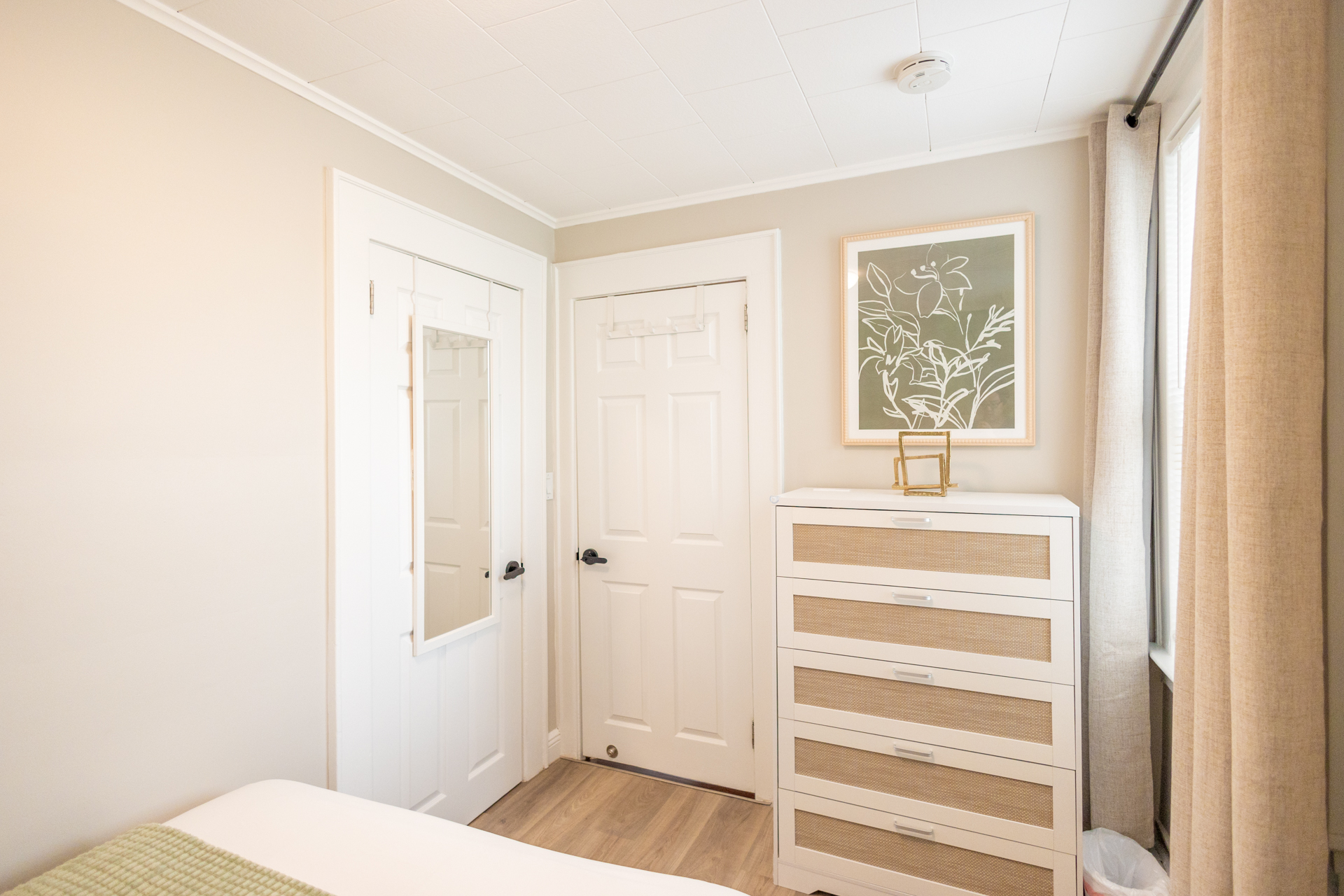 A white dresser with rattan-front drawers beside a closet door with full-length mirror, framed by tall curtains.