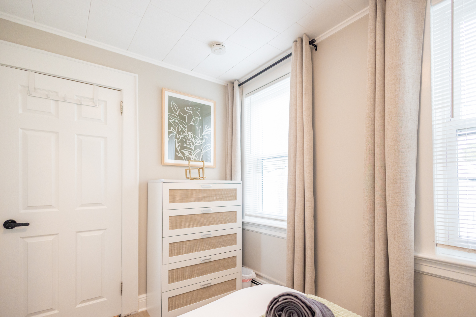 A white dresser with rattan drawers sits beneath a framed botanical print beside tall windows with beige curtains.