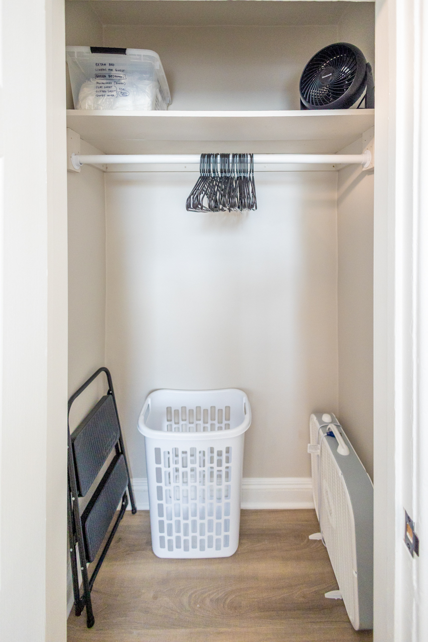 Open closet with hangers, a laundry basket, step stool, extra linens in a labeled bin, and a small fan on the shelf.