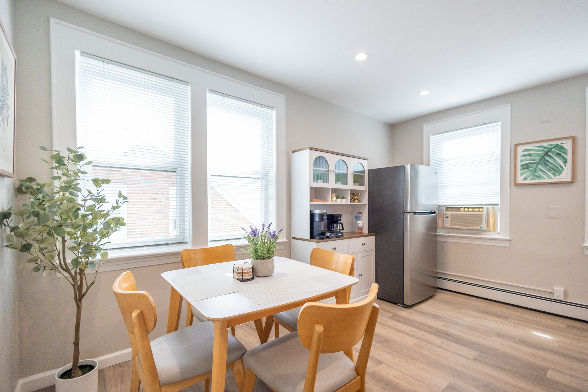 A bright dining nook with a wooden table and four chairs, a potted plant, large windows, and a hutch with a coffee maker next to the fridge.