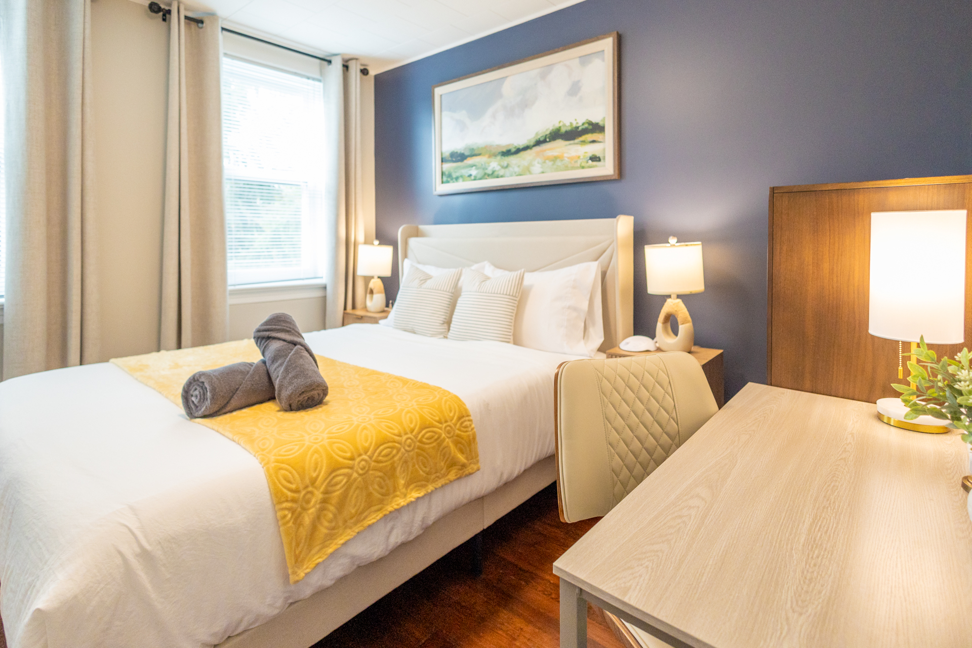 A cozy bedroom with a queen bed dressed in white linens, yellow blanket, and striped pillows. A wooden desk with a lamp and chair sits beside the bed, against a deep blue accent wall.