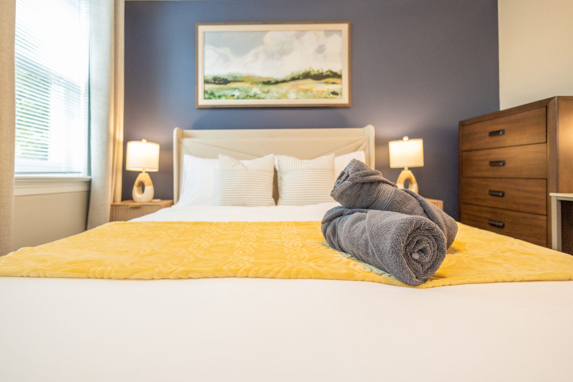 Close-up view of a queen bed with white linens, yellow blanket, striped pillows, and rolled gray towels on top. A dresser and lamps flank the bed.