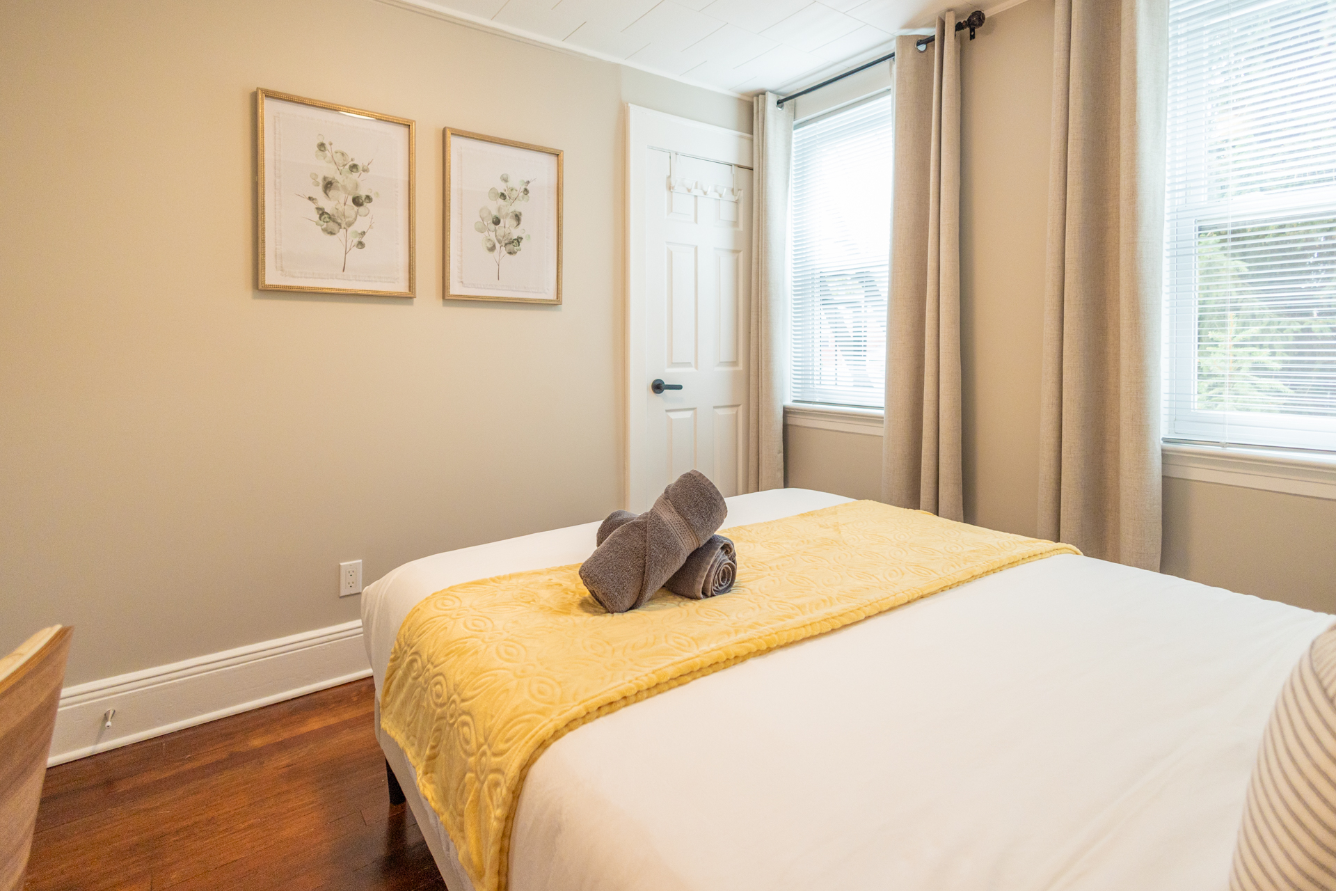 A queen bed with yellow blanket and striped pillows, desk and chair in the background, with framed artwork above the wall.