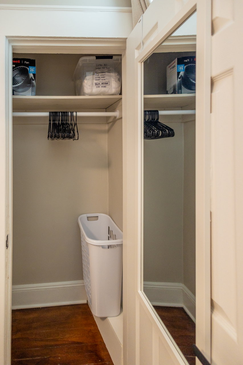 Open closet with laundry basket, hangers, extra bedding in a labeled bin, and a fan stored on the top shelf. A full-length mirror is mounted on the door.