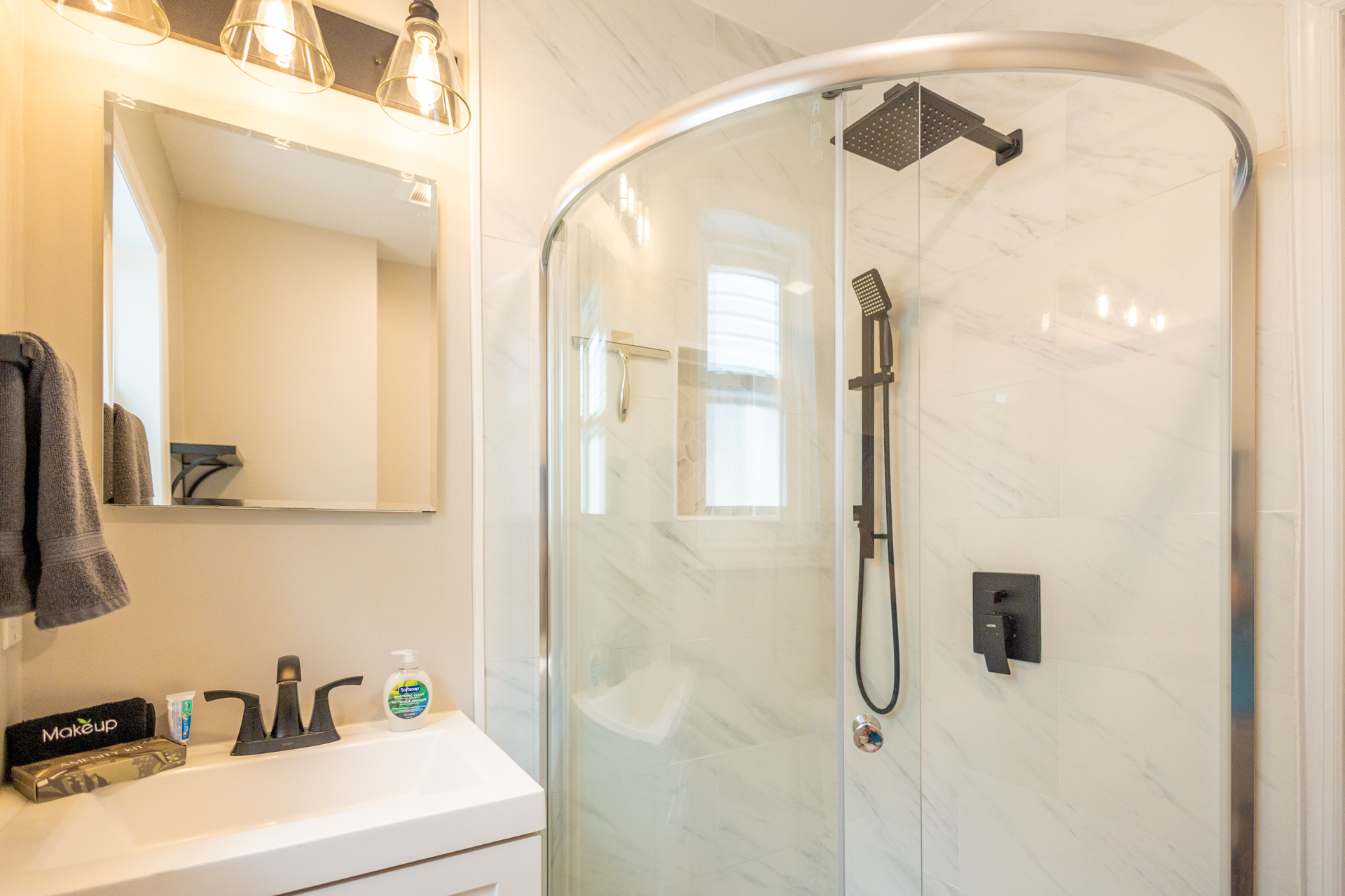 A modern bathroom with a glass-enclosed corner shower, rainfall showerhead, white vanity, and mirror with industrial light fixtures.