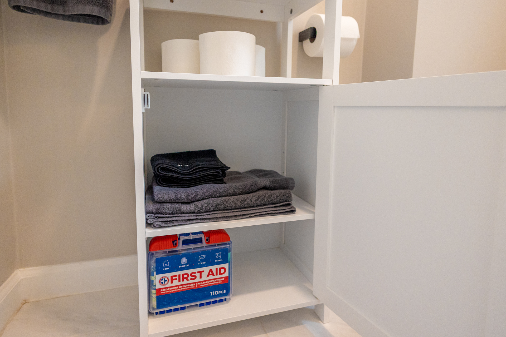 Open cabinet with folded gray towels, rolls of toilet paper, and a first aid kit.