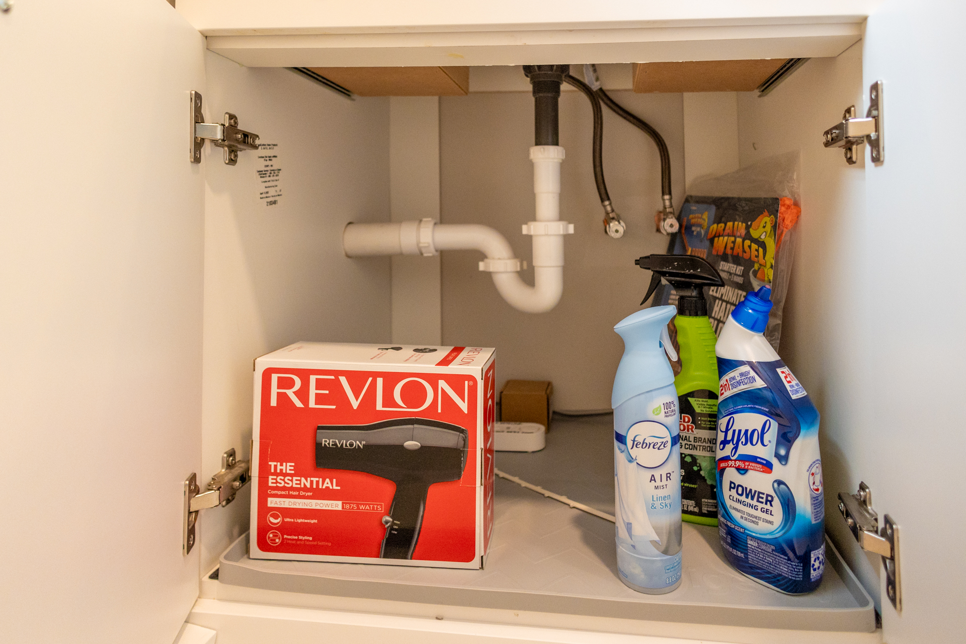 Open cabinet under bathroom sink with a Revlon hair dryer in box, Febreze spray, Lysol cleaner, and Drain Weasel cleaning supplies.