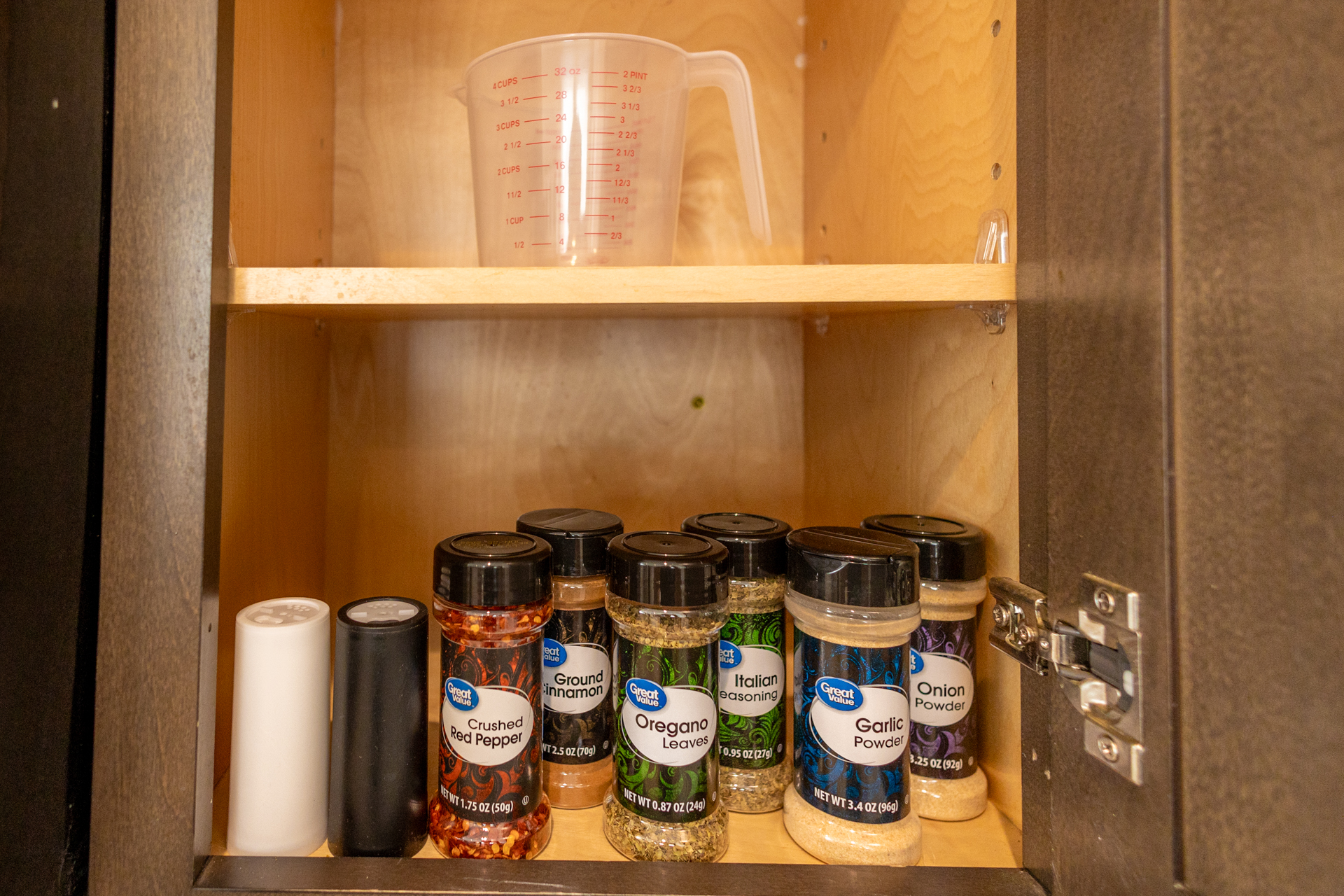 Open cabinet with measuring cup and a row of spices including oregano, Italian seasoning, garlic powder, and crushed red pepper.