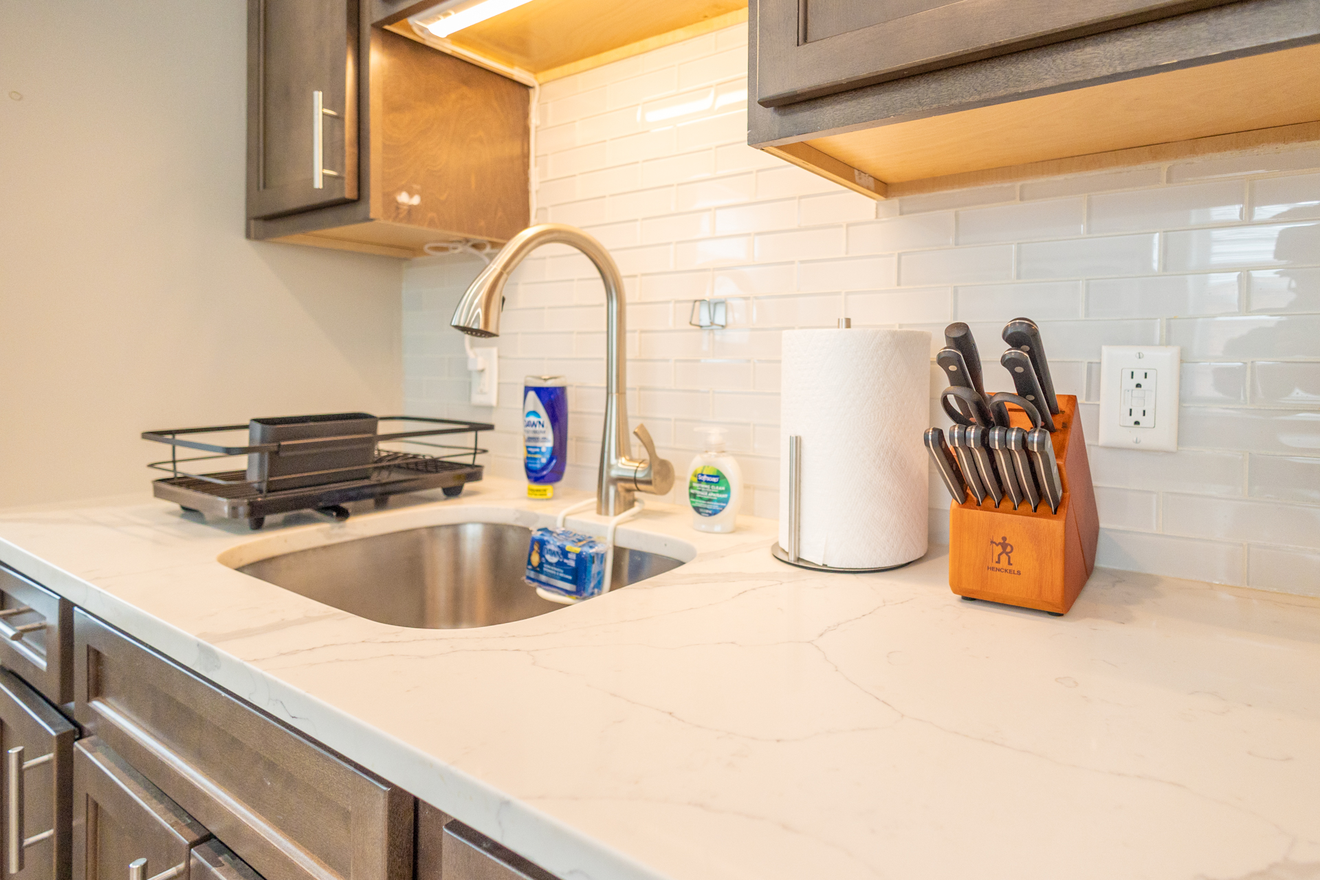 Kitchen sink with drying rack, paper towel holder, soap, and a knife block on a white marble counter.