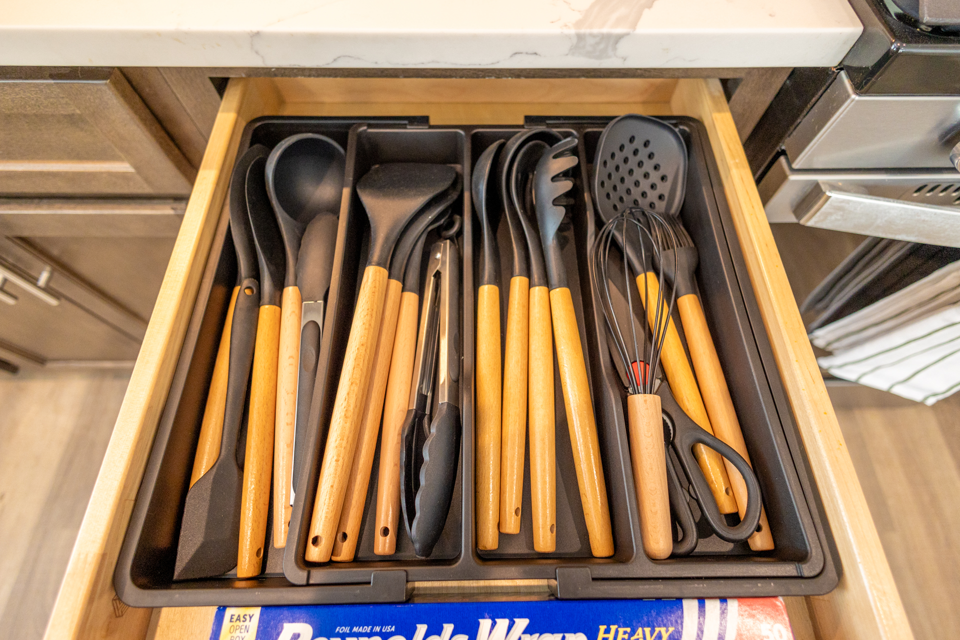 Drawer filled with wooden-handled black cooking utensils including spatulas, tongs, spoons, and a whisk.