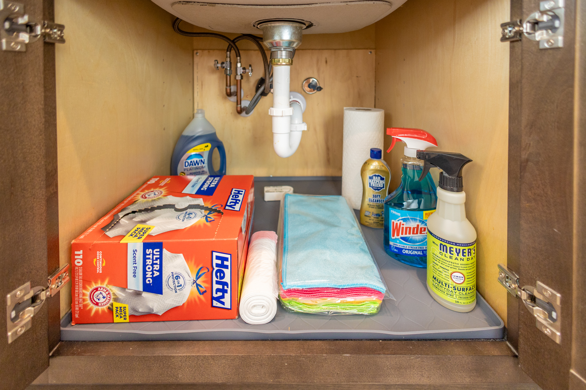 Open cabinet under the kitchen sink with Hefty trash bags, Dawn dish soap, microfiber cloths, Windex, Meyers spray, Bar Keeper’s Friend, and paper towels.