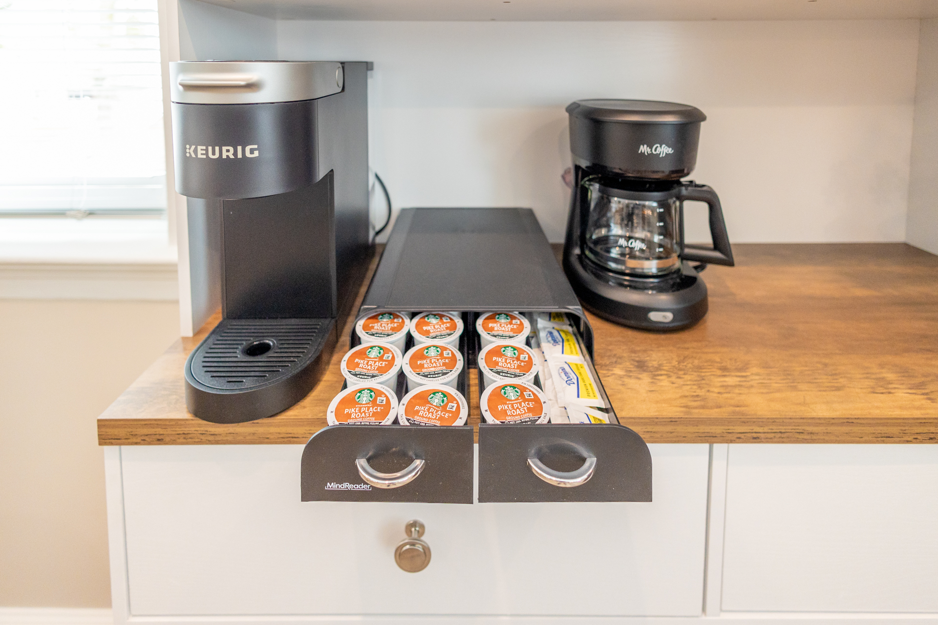 A Keurig coffee maker, a Mr. Coffee machine, and a drawer with Starbucks pods and sugar packets on a wooden counter.