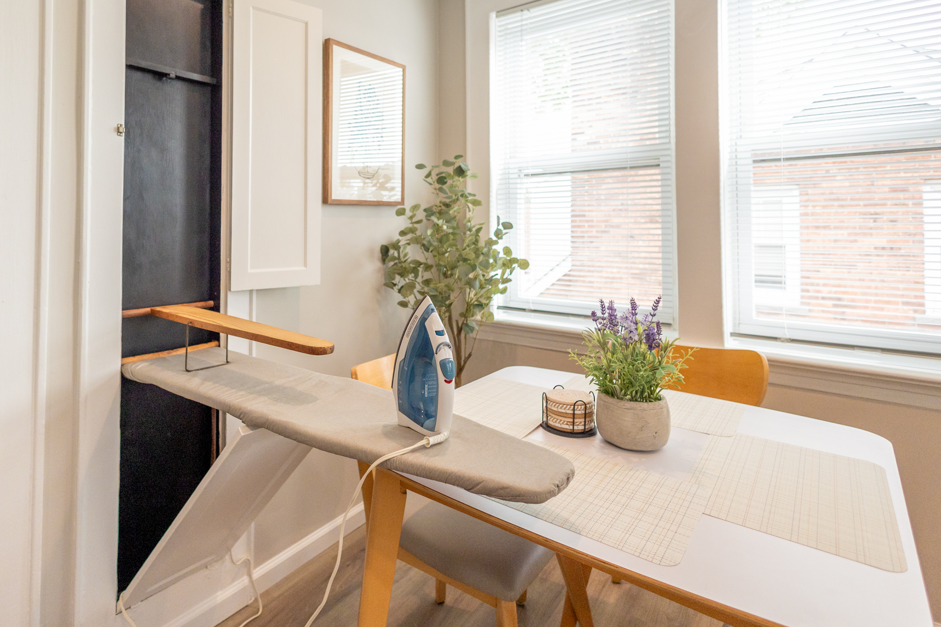 A wall-mounted pull-out ironing board with an iron set up next to a dining table and window.