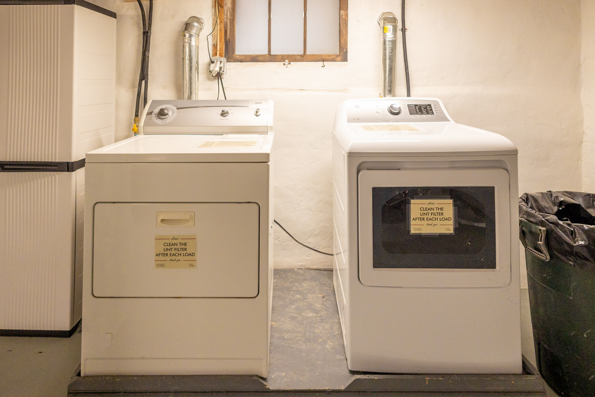Basement laundry area with white washer and dryer side by side, labeled with instructions.