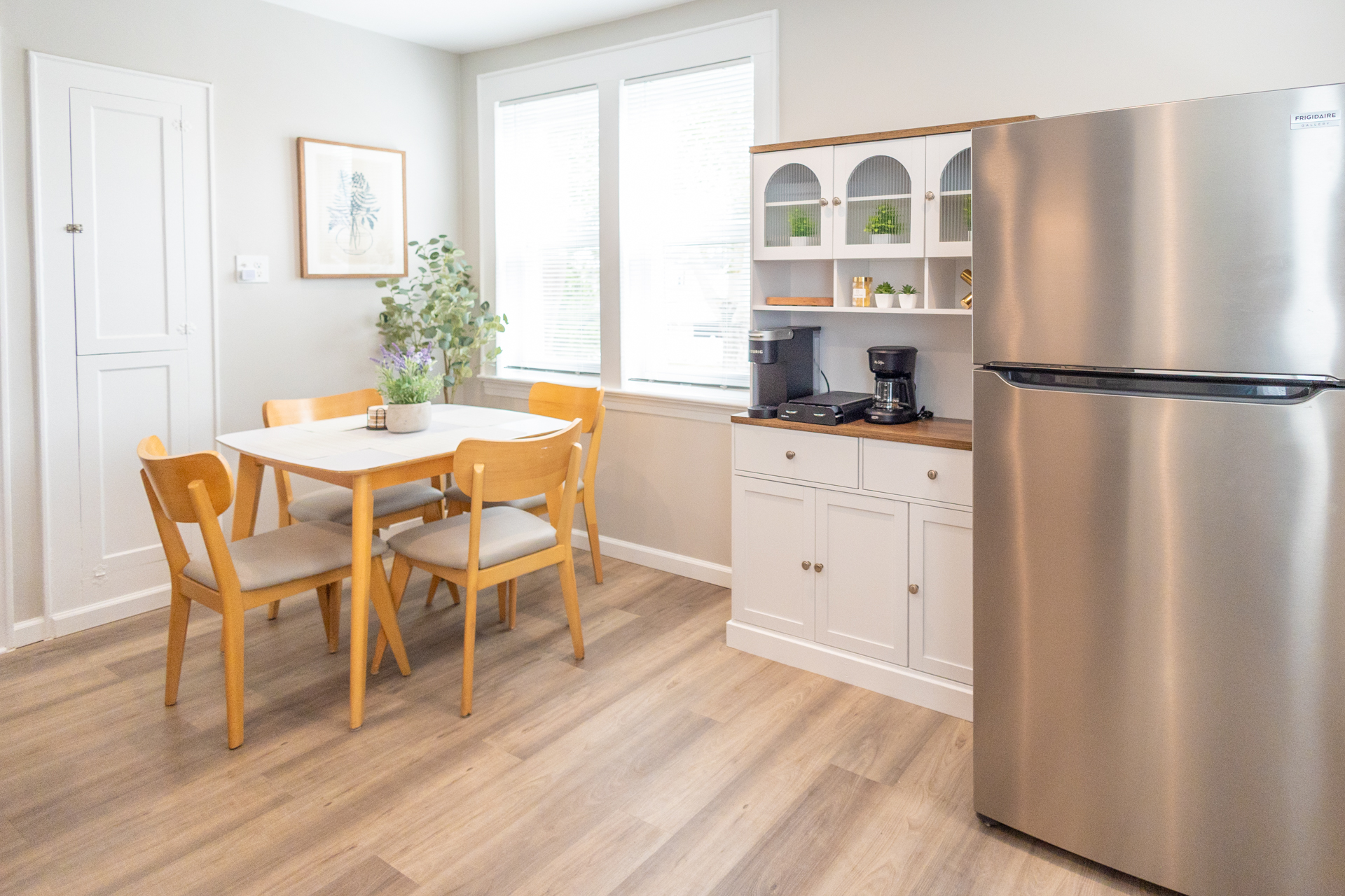 A light wooden dining table with four chairs sits beside a white hutch with coffee makers, next to a stainless steel fridge. Large windows let in natural light.