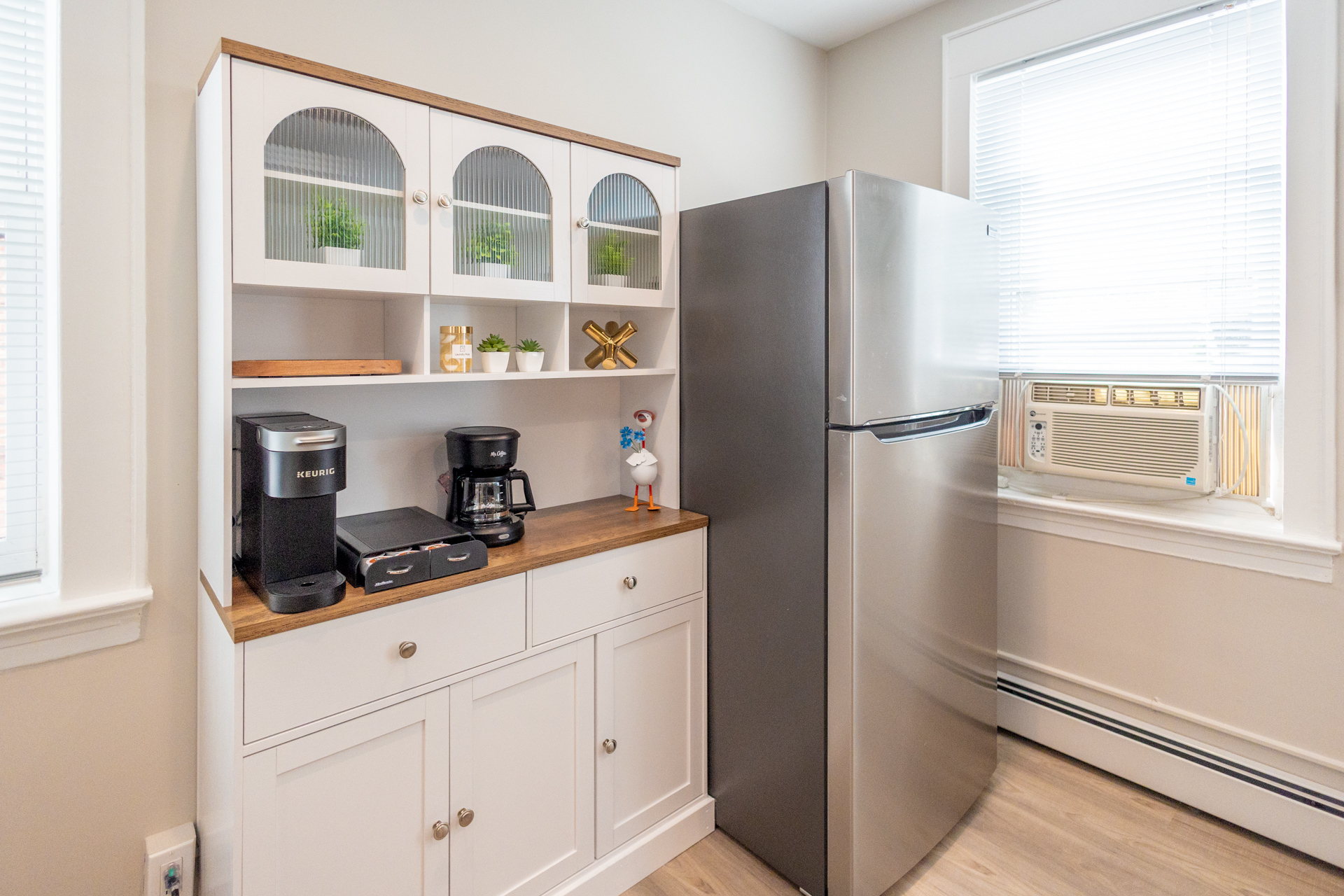 A white hutch with coffee makers and decorative accents sits next to a stainless steel fridge and window with an air conditioner.