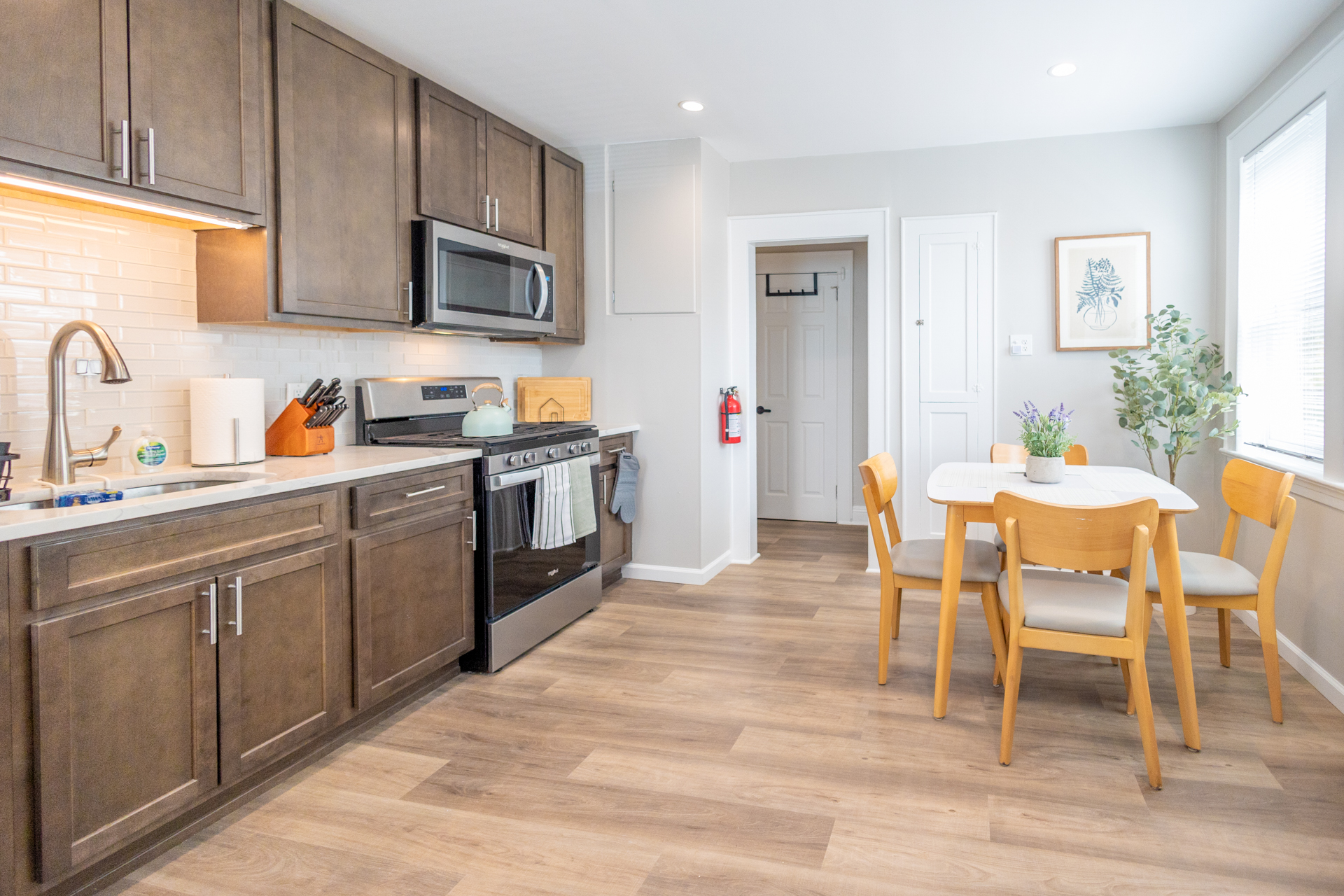 A modern kitchen with dark wood cabinets, stainless steel stove and microwave, and a dining table for four beside a large window.