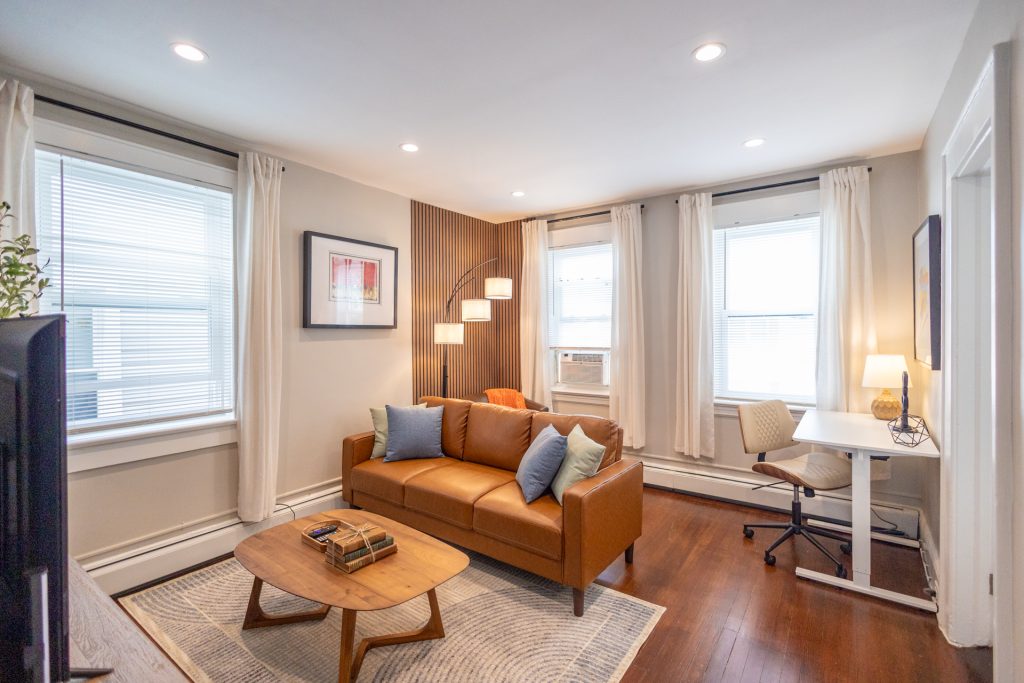 Wide view of the living room with a leather sofa, modern floor lamp, small desk, and large windows with white curtains. A coffee table sits on a patterned rug in the center.