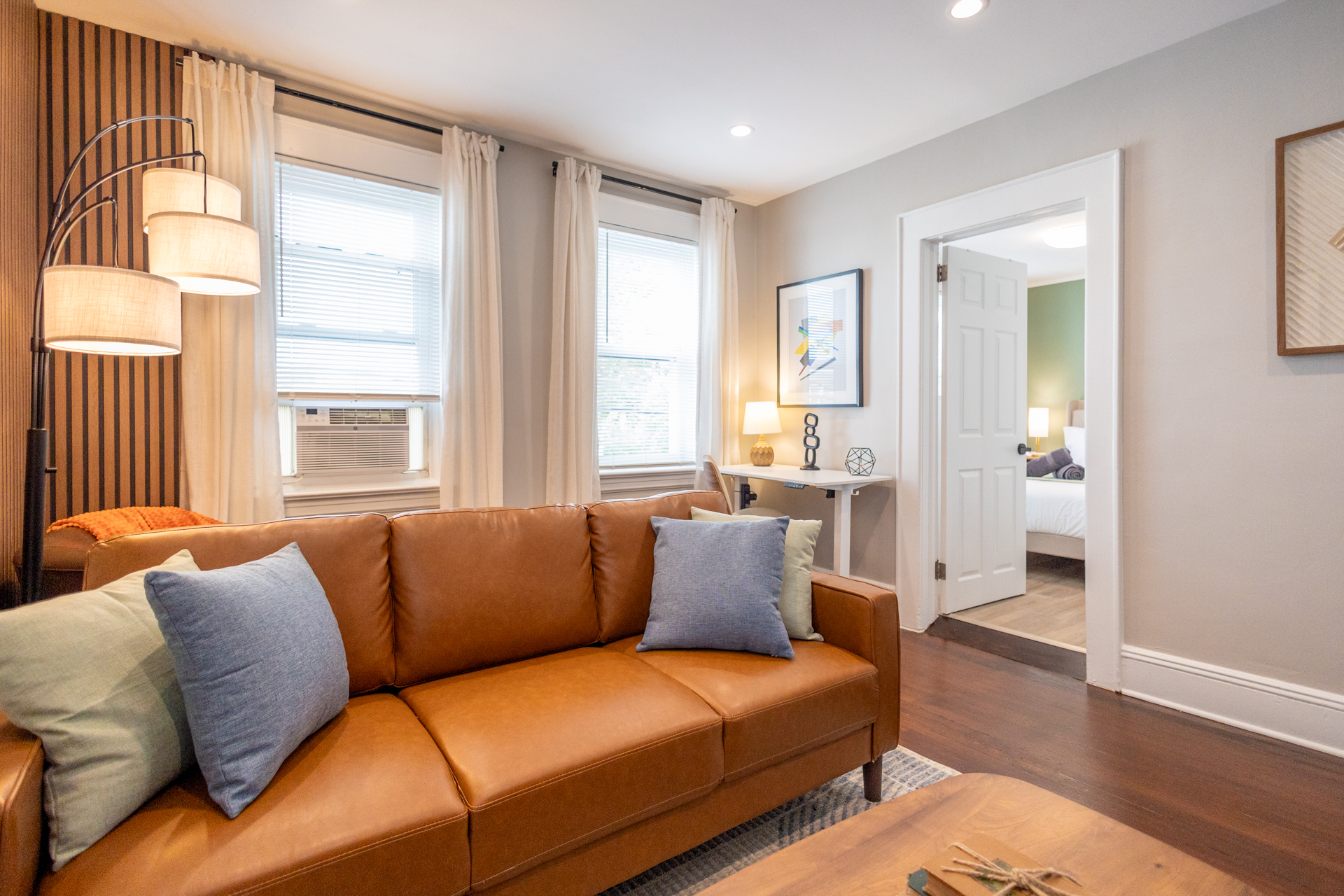A brown leather sofa with colorful pillows sits in front of tall windows with white curtains. A desk with a lamp and chair is placed in the corner, near the entrance to the bedroom.