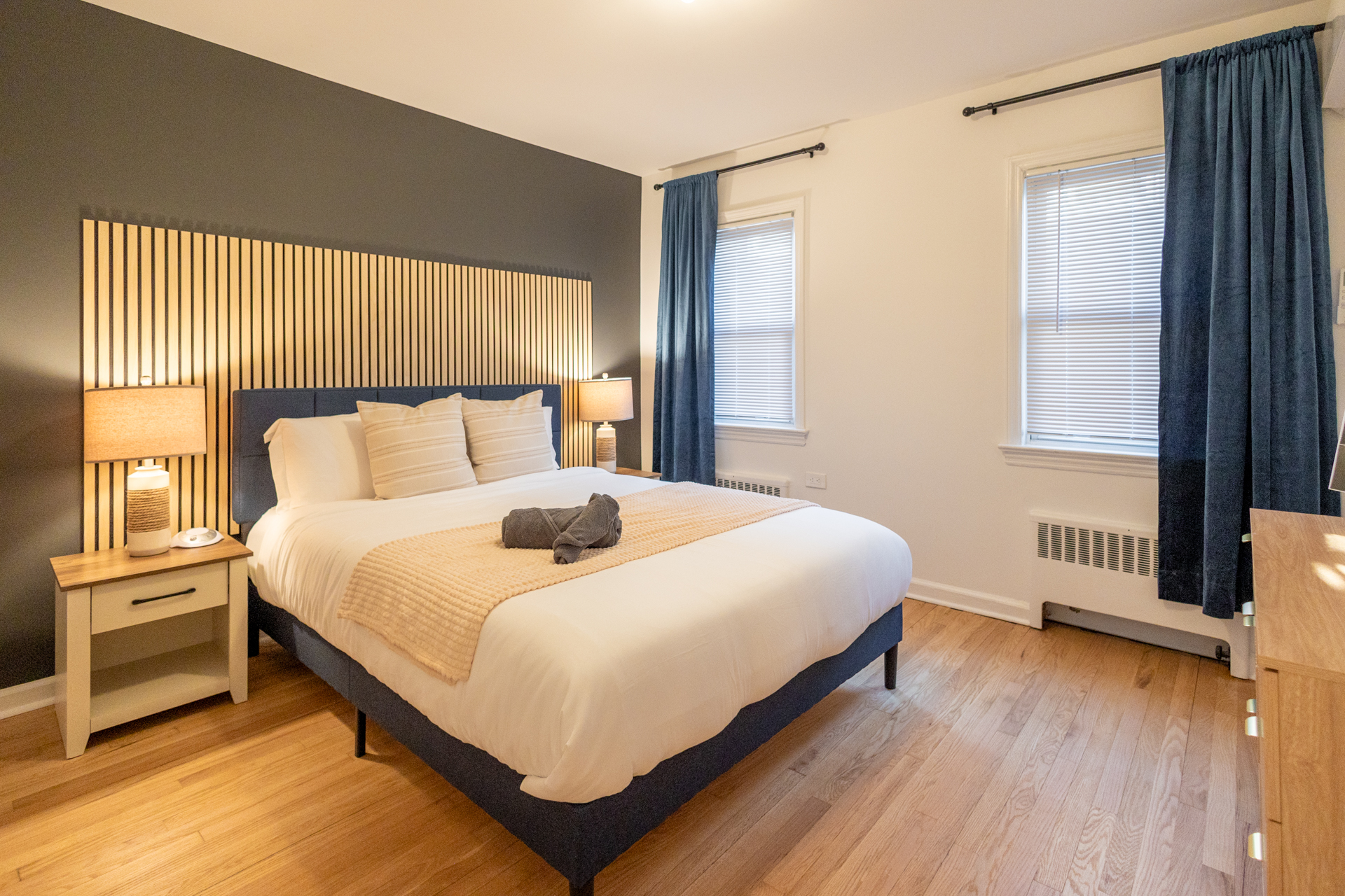 Featuring a navy blue fabric frame with neatly made with crisp white linens, a soft peach-colored textured throw blanket placed at the foot, and two gray rolled towels on top.