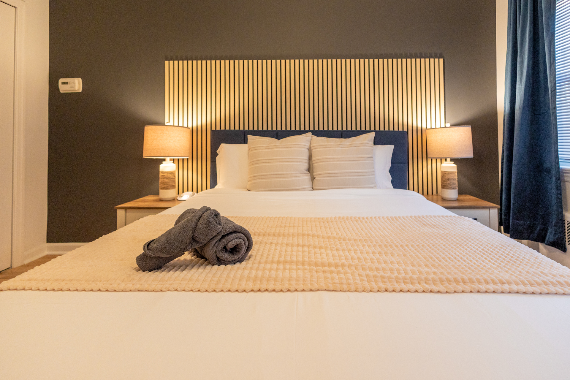 Cozy bedroom with a dark wood and navy headboard, cream pillows, and an orange throw, framed by matching nightstands and warm bedside lamps.