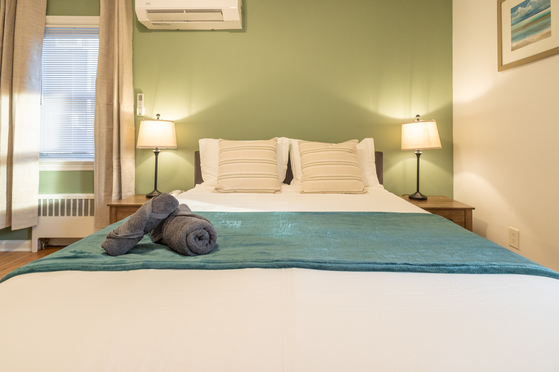 Queen bedroom with white bedding, teal blanket, sage wall, and soft lamp light.