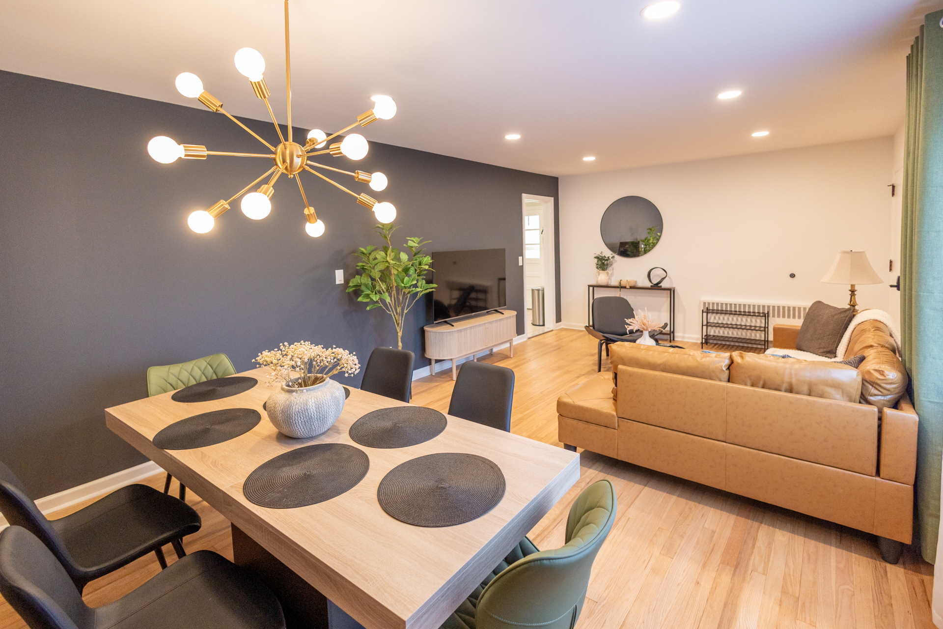 A modern, open-concept dining and living space featuring warm wood tones, a bold charcoal accent wall, a gold sputnik chandelier, and a cozy caramel sofa, all styled with clean lines and soft decorative touches.