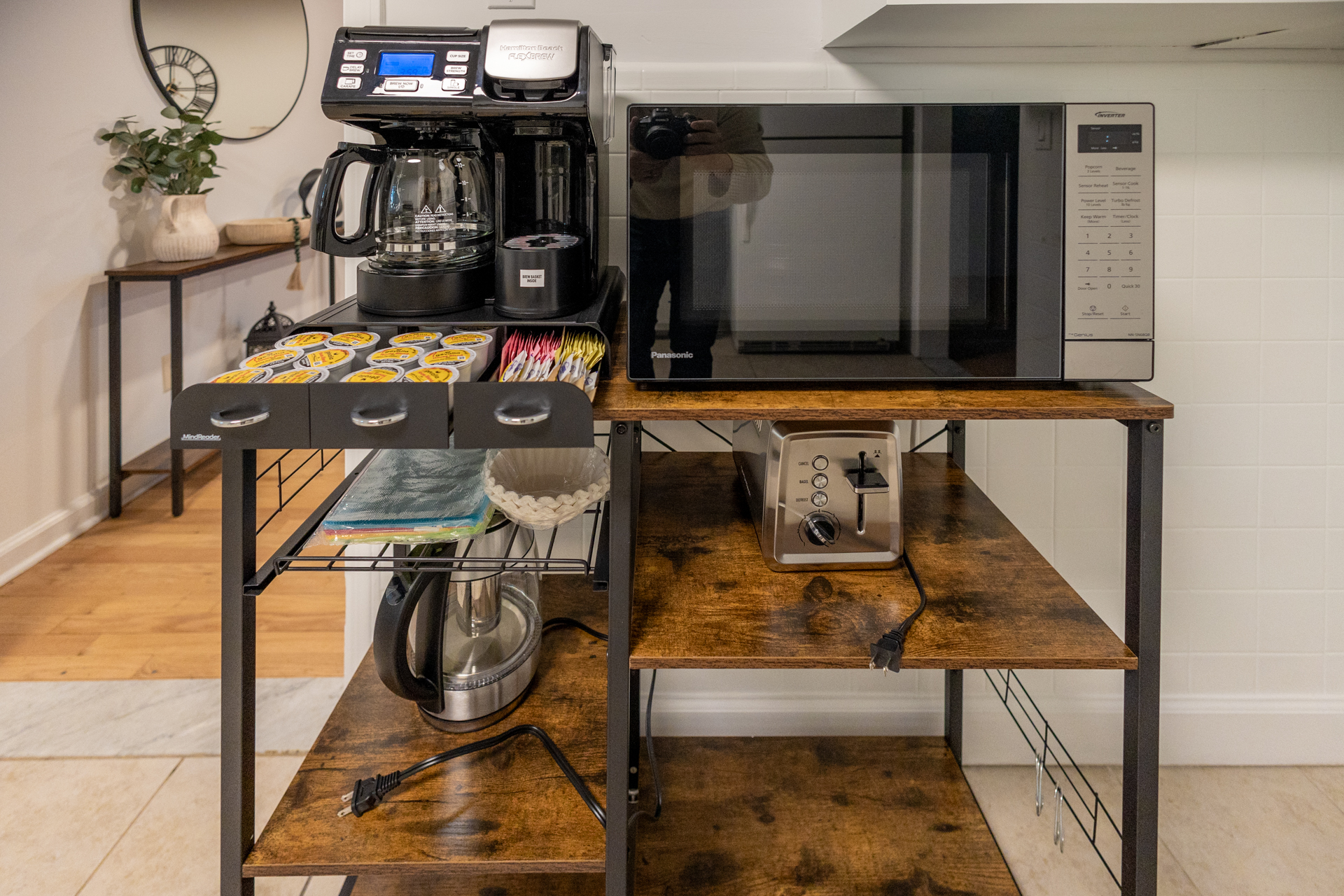 Coffee and breakfast station with a drip coffee maker, K-cups, microwave, toaster, and electric kettle arranged neatly on a wooden metal rack.