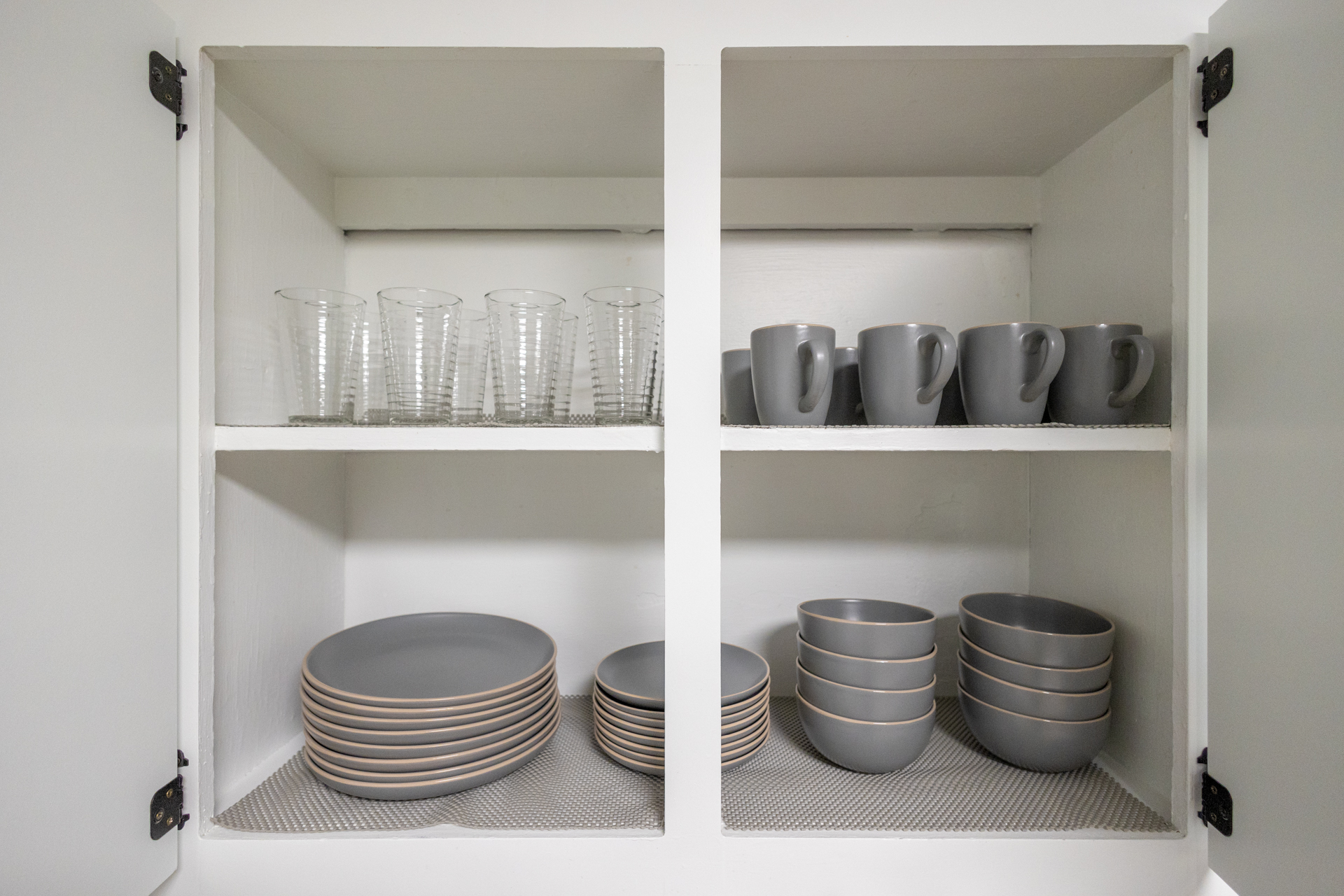 Kitchen cabinet neatly stocked with glassware, grey mugs, plates, and bowls