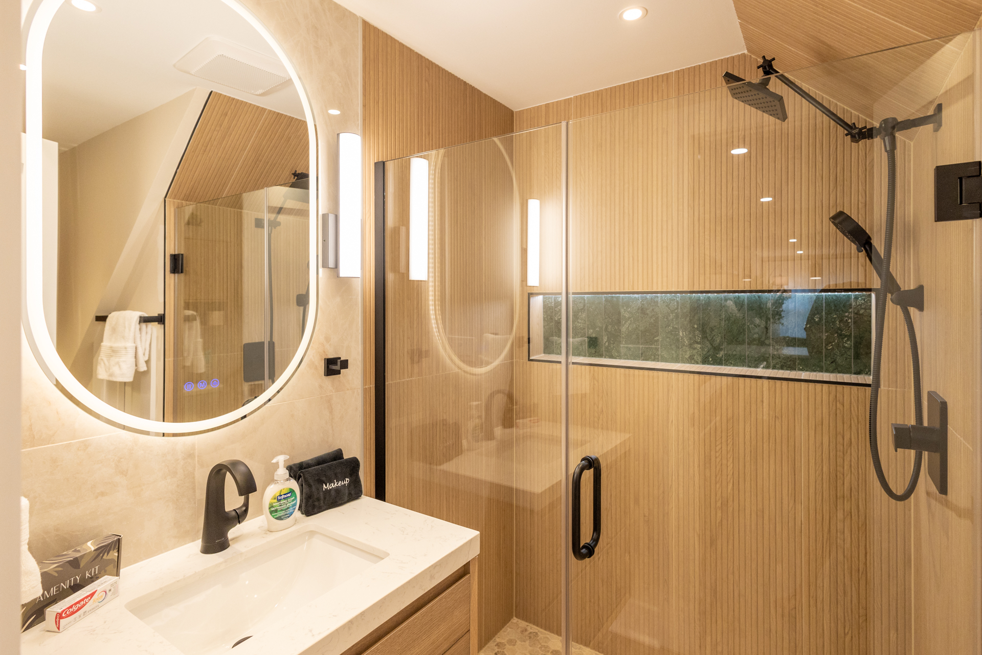 Featuring a white vanity, illuminated mirror, and glass-enclosed walk-in shower with dual showerheads.
